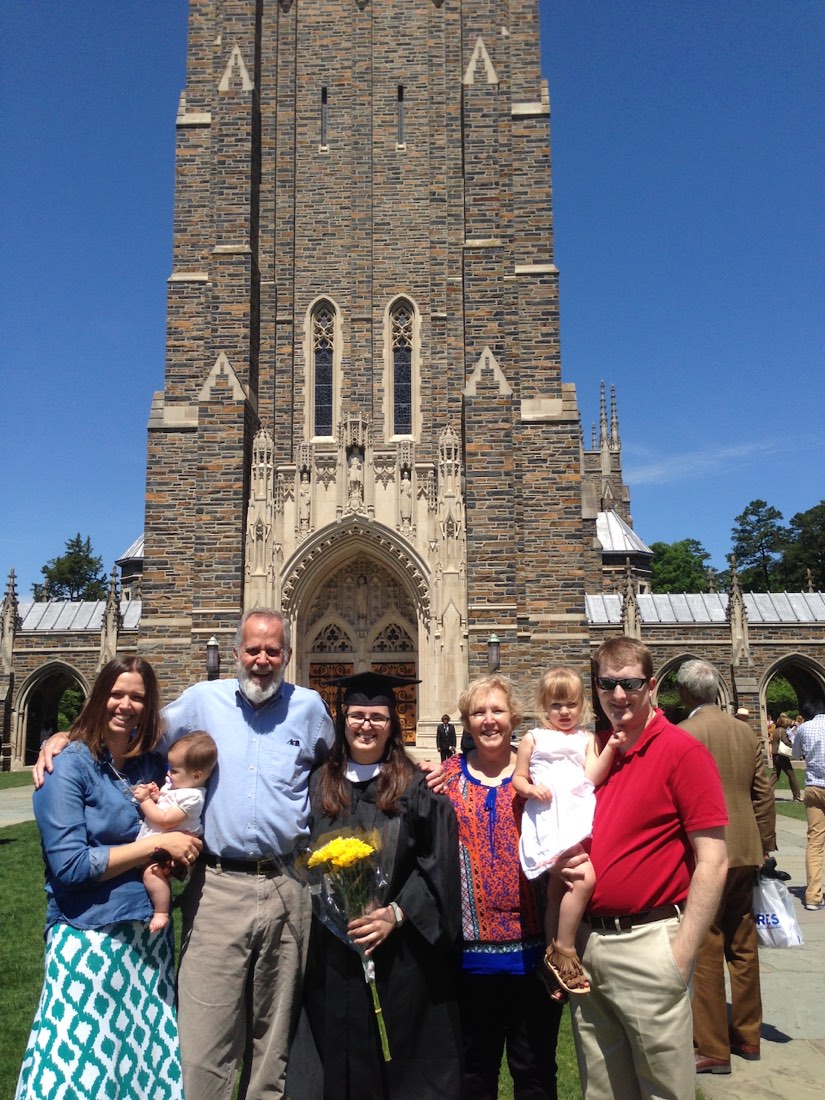 Maggie in a cap and gown, holding yellow flowers, with her family in front of Duke Chapel