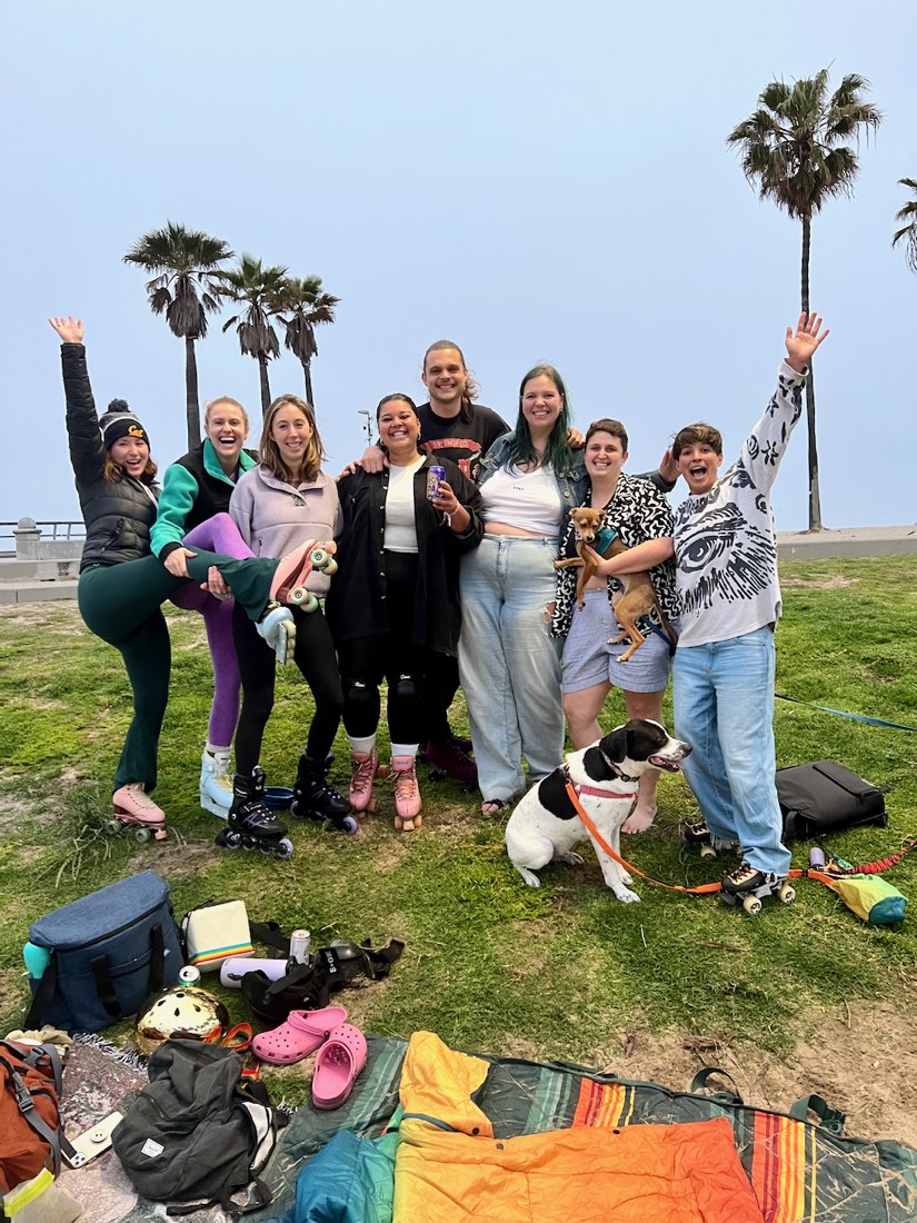 Maggie holding Brie, surrounded by friends in roller skates on the grass by the beach, with palm trees behind them