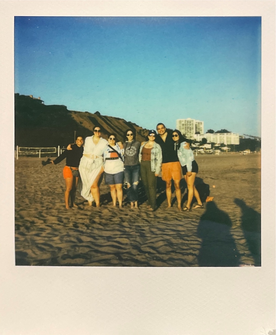 A Polaroid of seven friends standing arm in arm on the sand at golden hour
