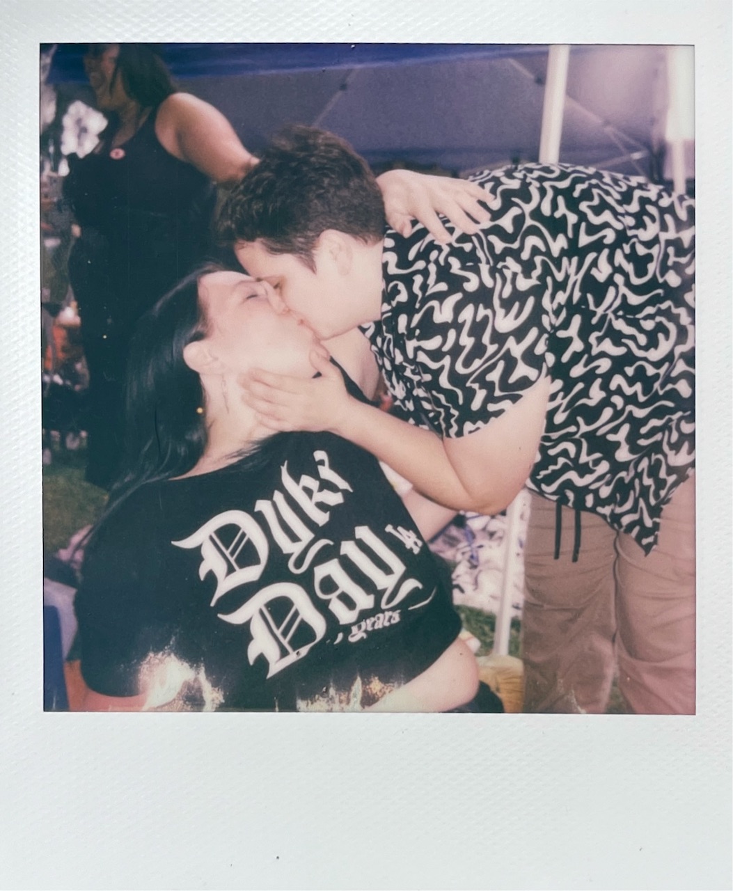 A Polaroid of Maggie leaning down to kiss Kristin, who's sitting in a Dyke Day shirt