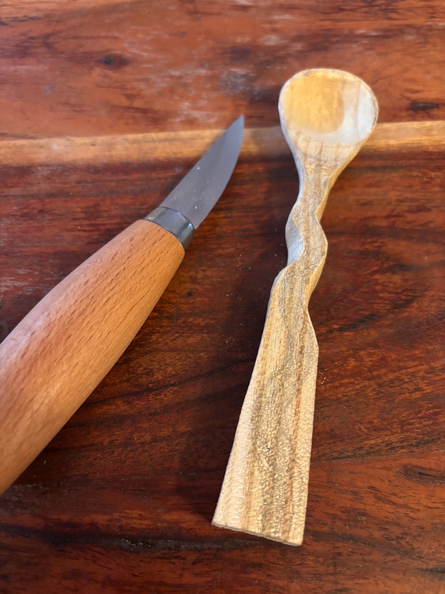 A pale spoon with a wavy, twisting handle lying next to a carving knife on a dark wood table