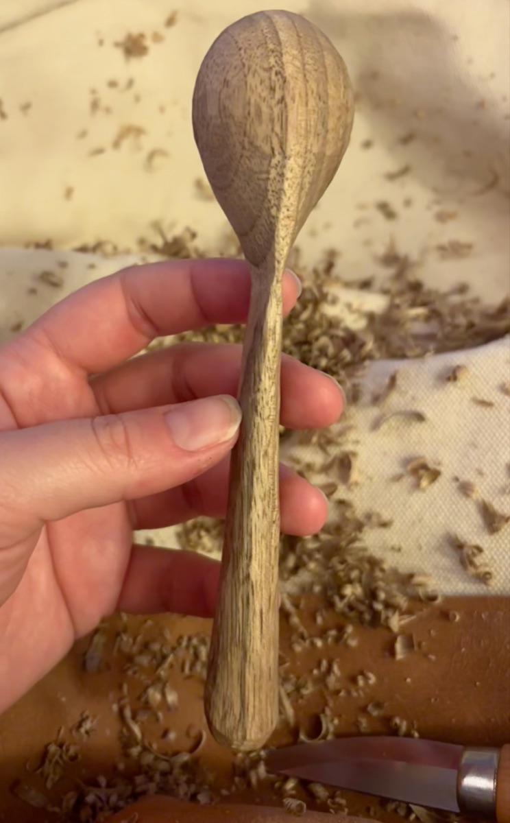 Maggie's hand holding a half-carved wooden spoon over a cloth covered in wood shavings, with a carving knife below