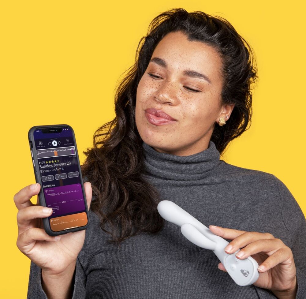 A woman against a yellow background holding a phone with the Lioness app's session charts in one hand and a white Lioness vibrator in the other
