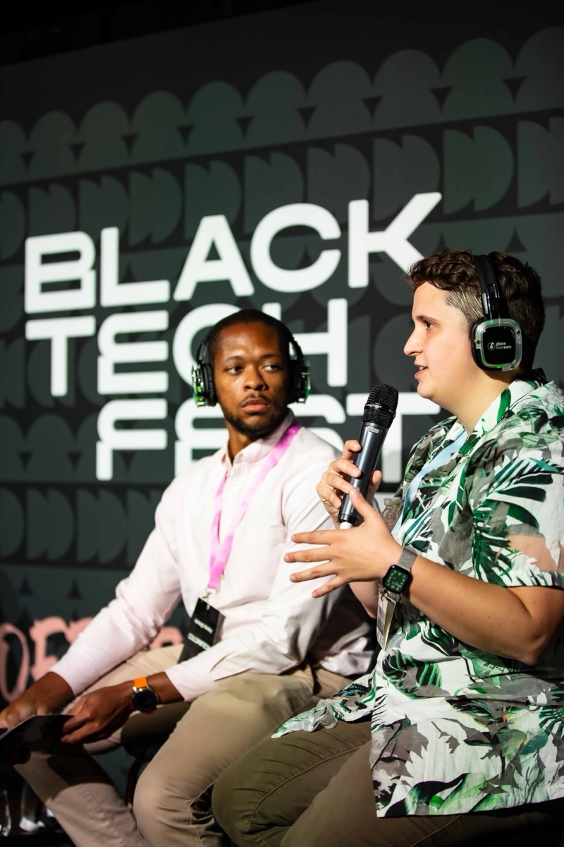Maggie on stage at Black Tech Fest in a green and white tropical-print shirt and headphones, speaking into a microphone next to another panelist, with the Black Tech Fest logo behind them